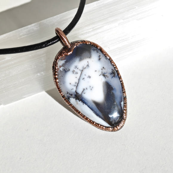 Colgante de merlinita —ópalo dendrítico— en bisel de cobre electroformado. Patrón de dendrites negras sobre fondo blanco y gris.